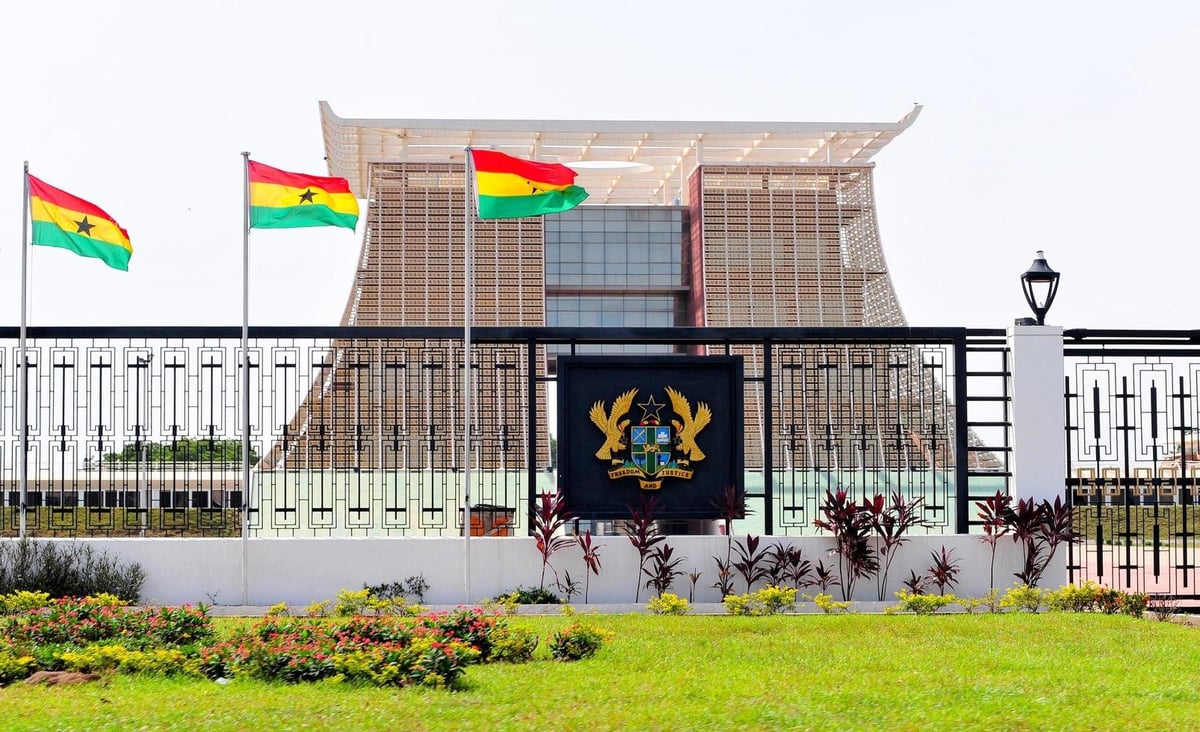 Modern government building with Ghanaian flags and national coat of arms displayed on facade, surrounded by green lawn and metal fencing